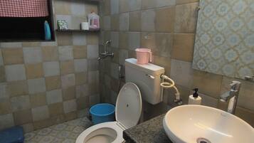 Une salle de bain avec des toilettes, un lavabo et une douche. On y trouve des étagères avec des articles de toilette et une fenêtre avec un store à carreaux.