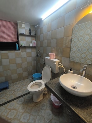 Une salle de bain avec des toilettes, un lavabo et une douche. On y trouve des étagères avec des articles de toilette et une fenêtre avec un store à carreaux.