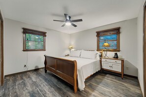 A bedroom with a wooden bed, a ceiling fan, two windows with blinds, a nightstand with a lamp, and a wall-mounted shelf with a decorative item. - Lovely 2-bedroom cabin in the Smoky Mountains in a gated community (Franklin)