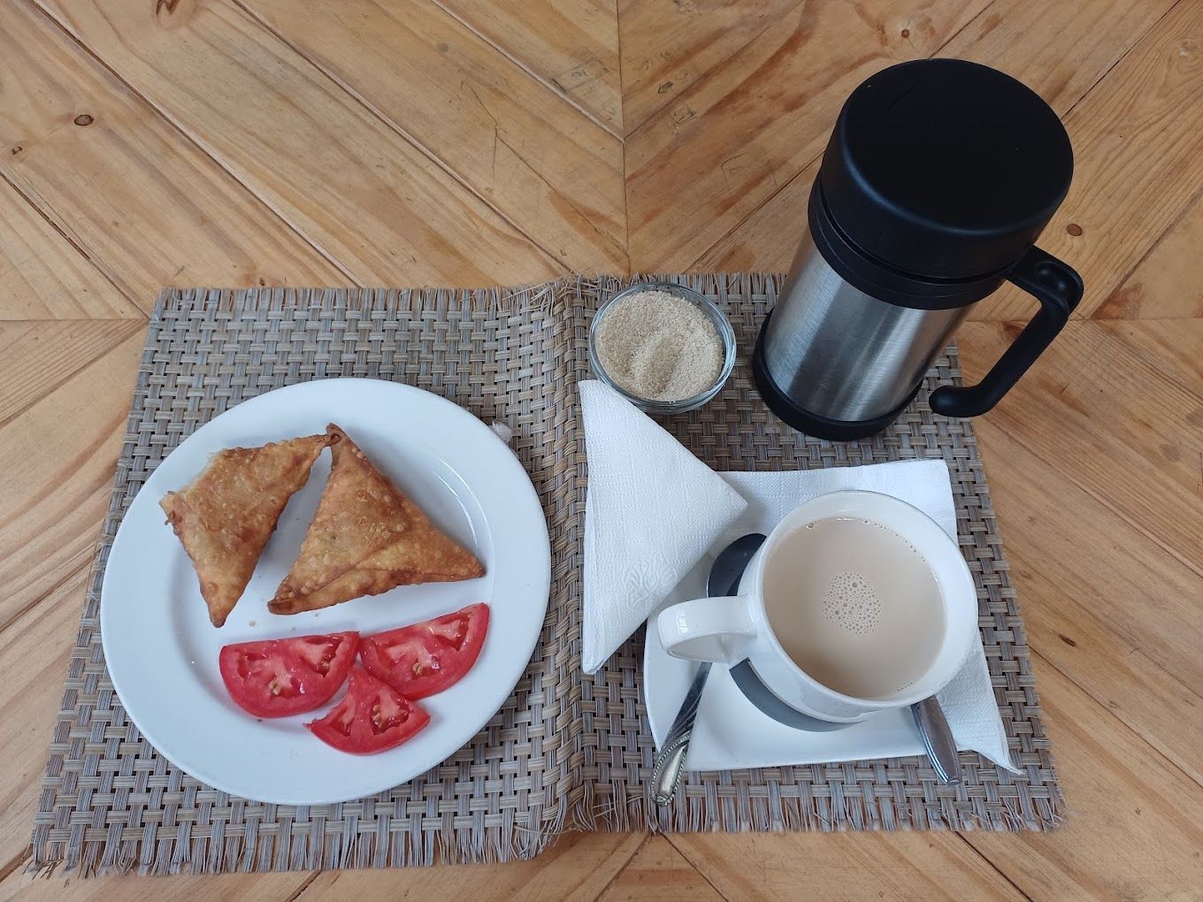 Un plato con dos porciones triangulares de comida y rodajas de tomate, una taza de café, un termo y un cuenco pequeño de azúcar sobre un camino de mesa tejido.