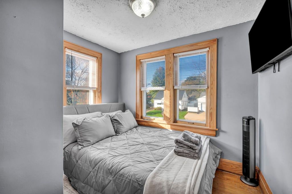 A bedroom with a bed, two windows with blinds, a ceiling light, and a black standing heater.