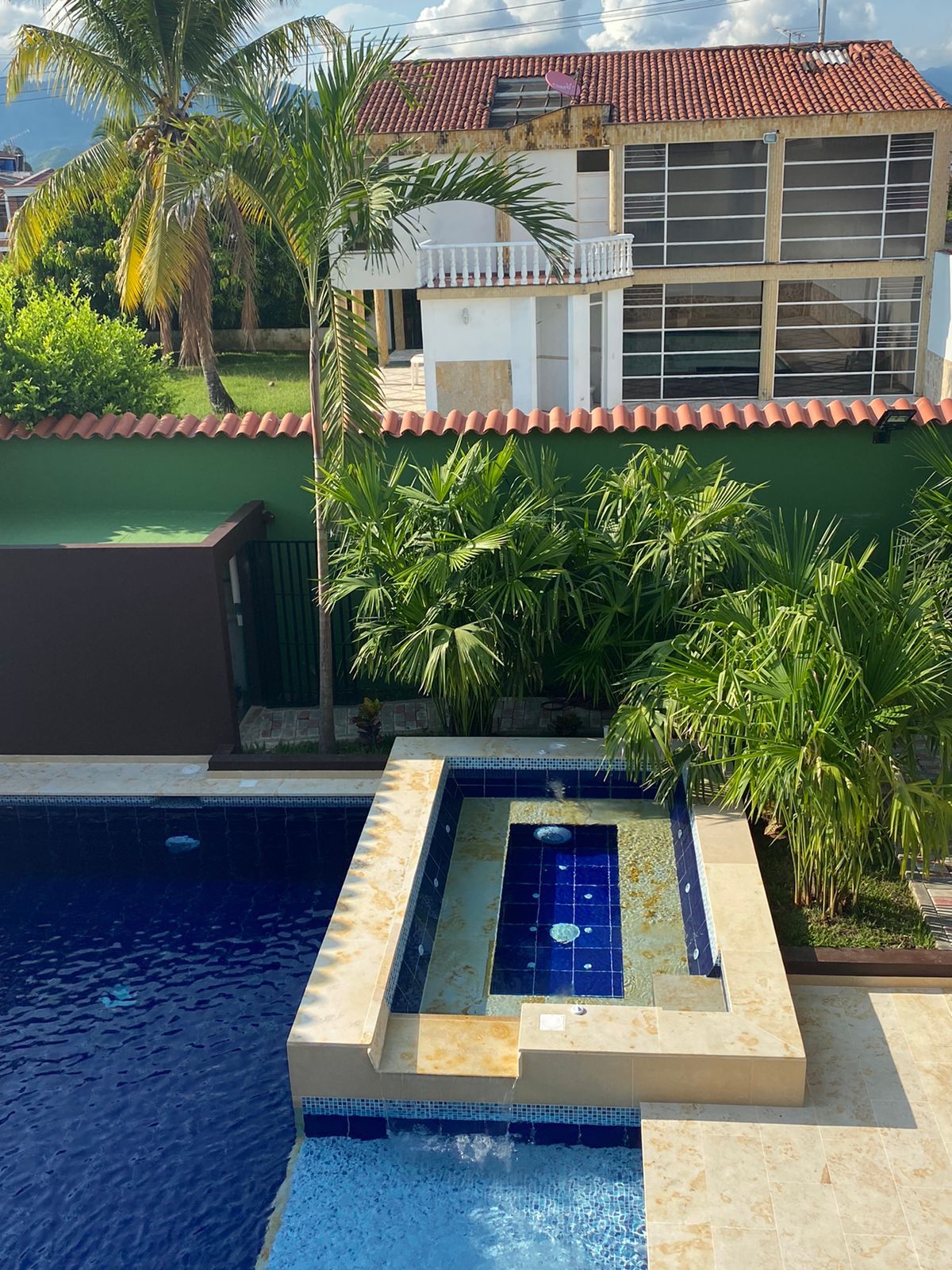 A swimming pool with a tiled deck, surrounded by lush greenery and a house with a red-tiled roof.