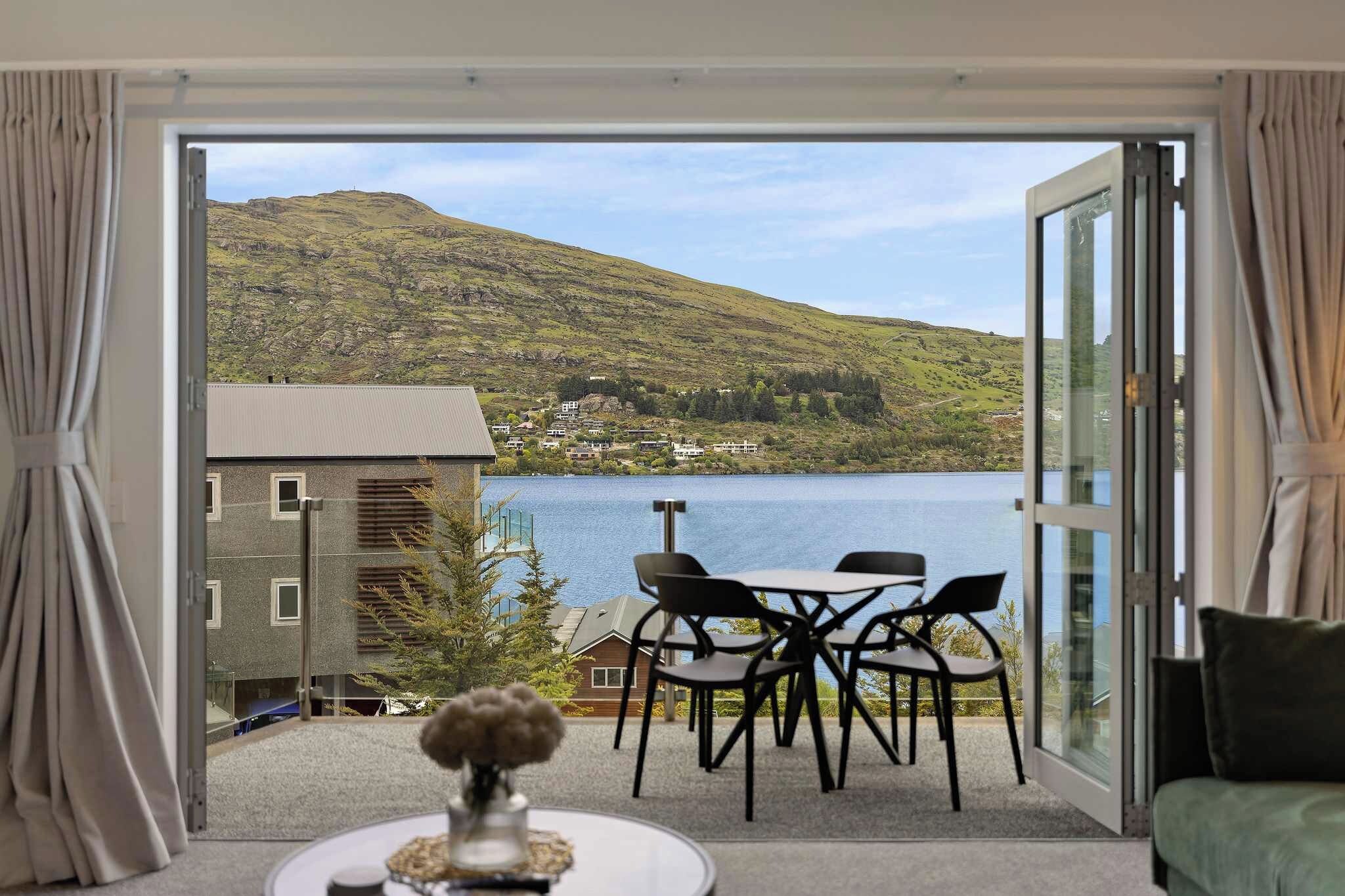 A room with a view of a lake and hills, a dining area with chairs and a table, and a sofa.