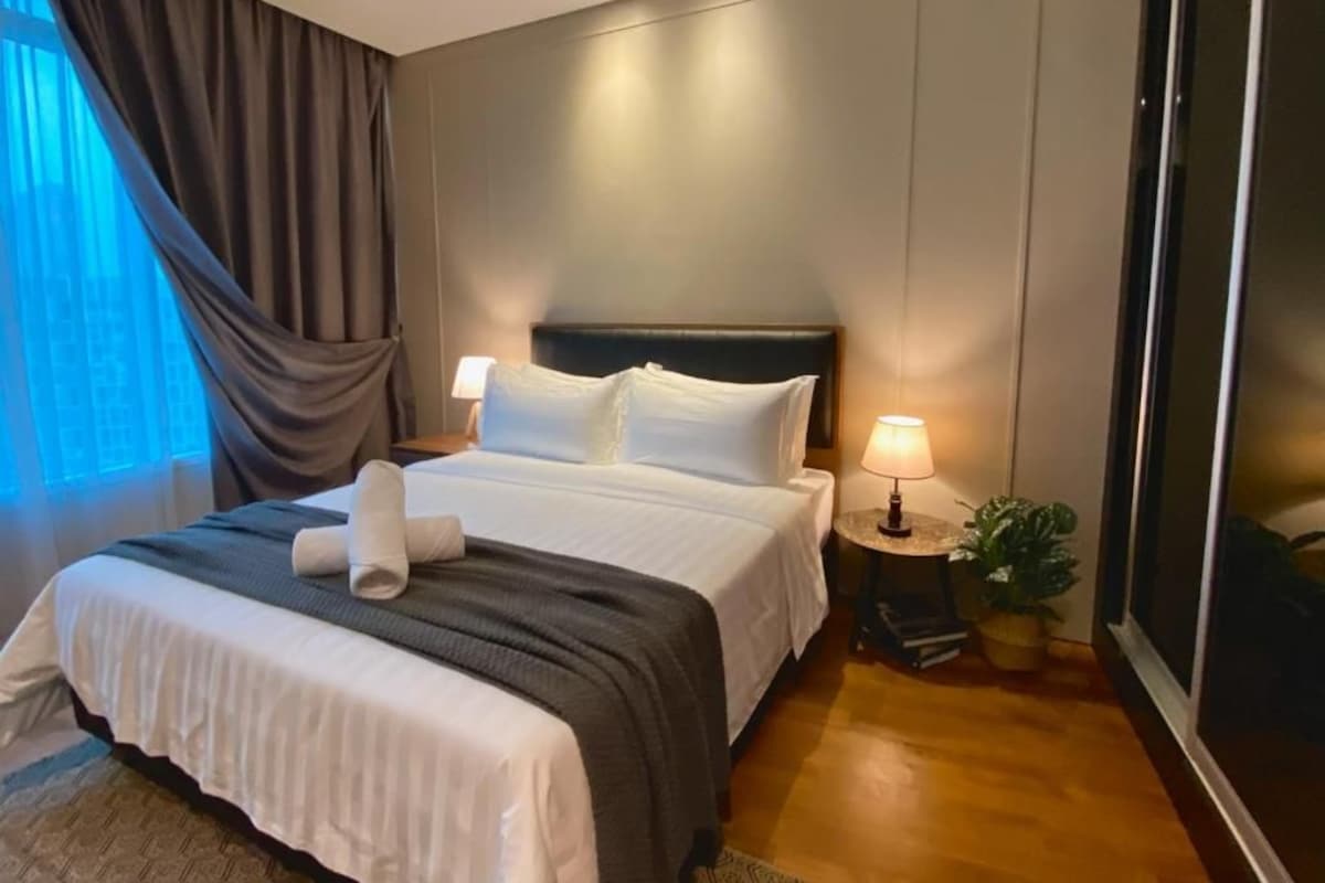 Habitación de hotel con una cama grande, una mesita de noche con lámpara y vistas a la ciudad desde la ventana.