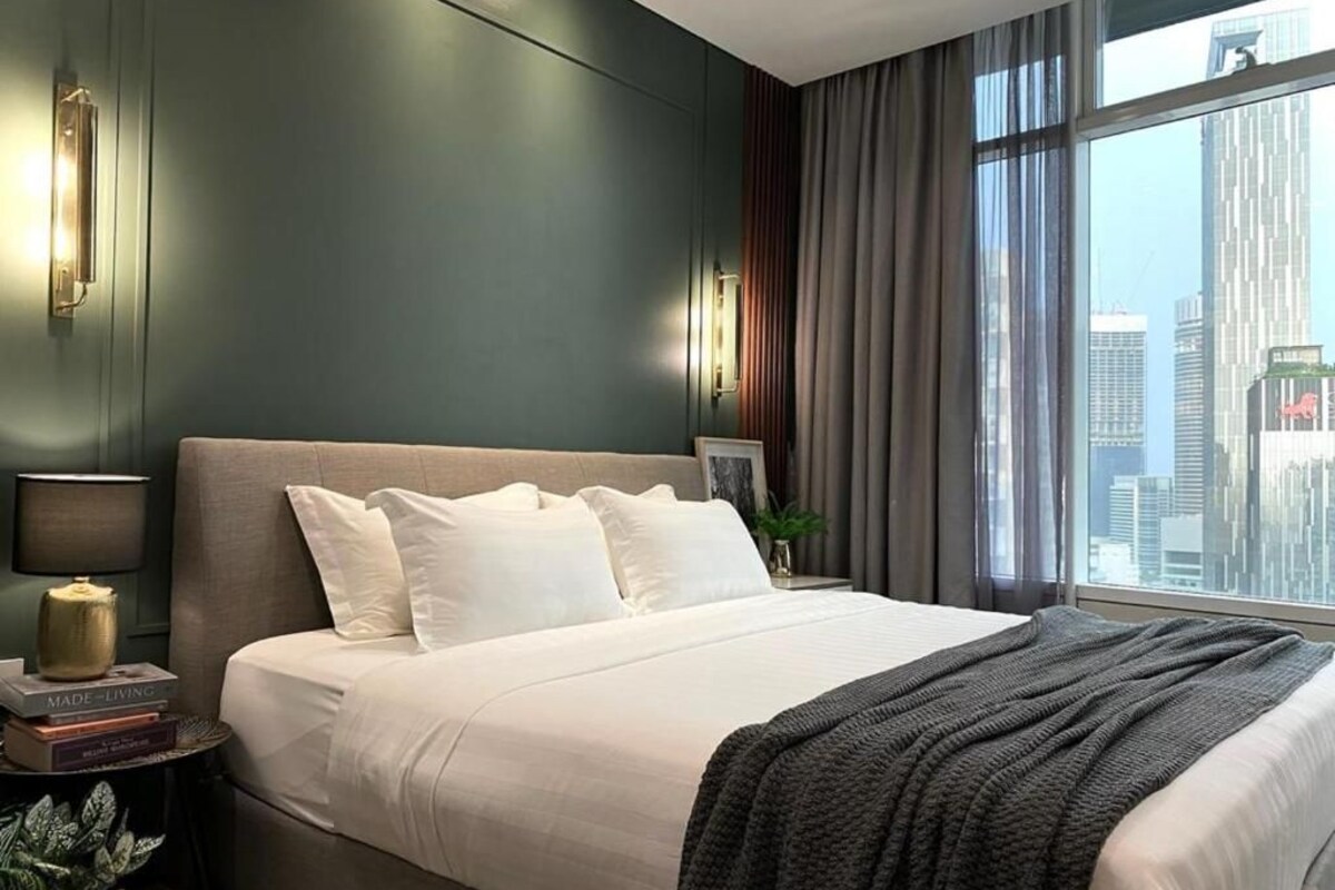 Habitación de hotel moderna con una cama grande, una mesita de noche, una lámpara y vistas a la ciudad a través de grandes ventanales.