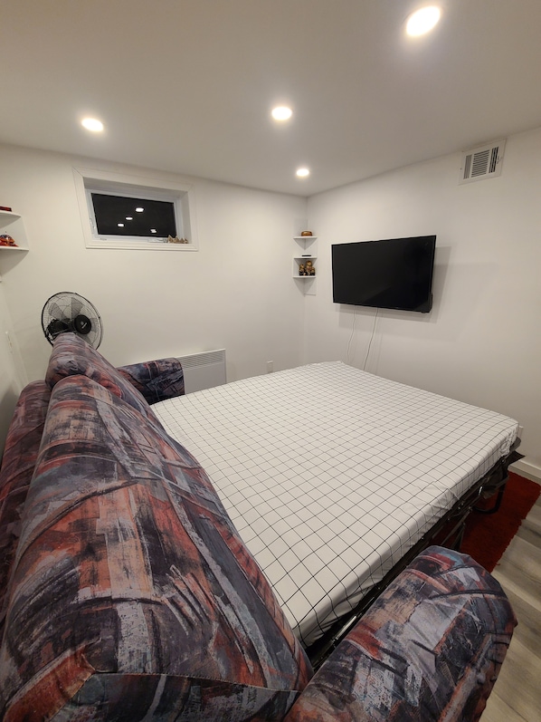 Un dormitorio con cama, televisor montado en la pared, ventilador de techo y ventana.