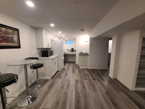 Una cocina moderna con armarios blancos, microondas y refrigerador. Hay un pequeño comedor con una mesa de vidrio y dos sillas.
