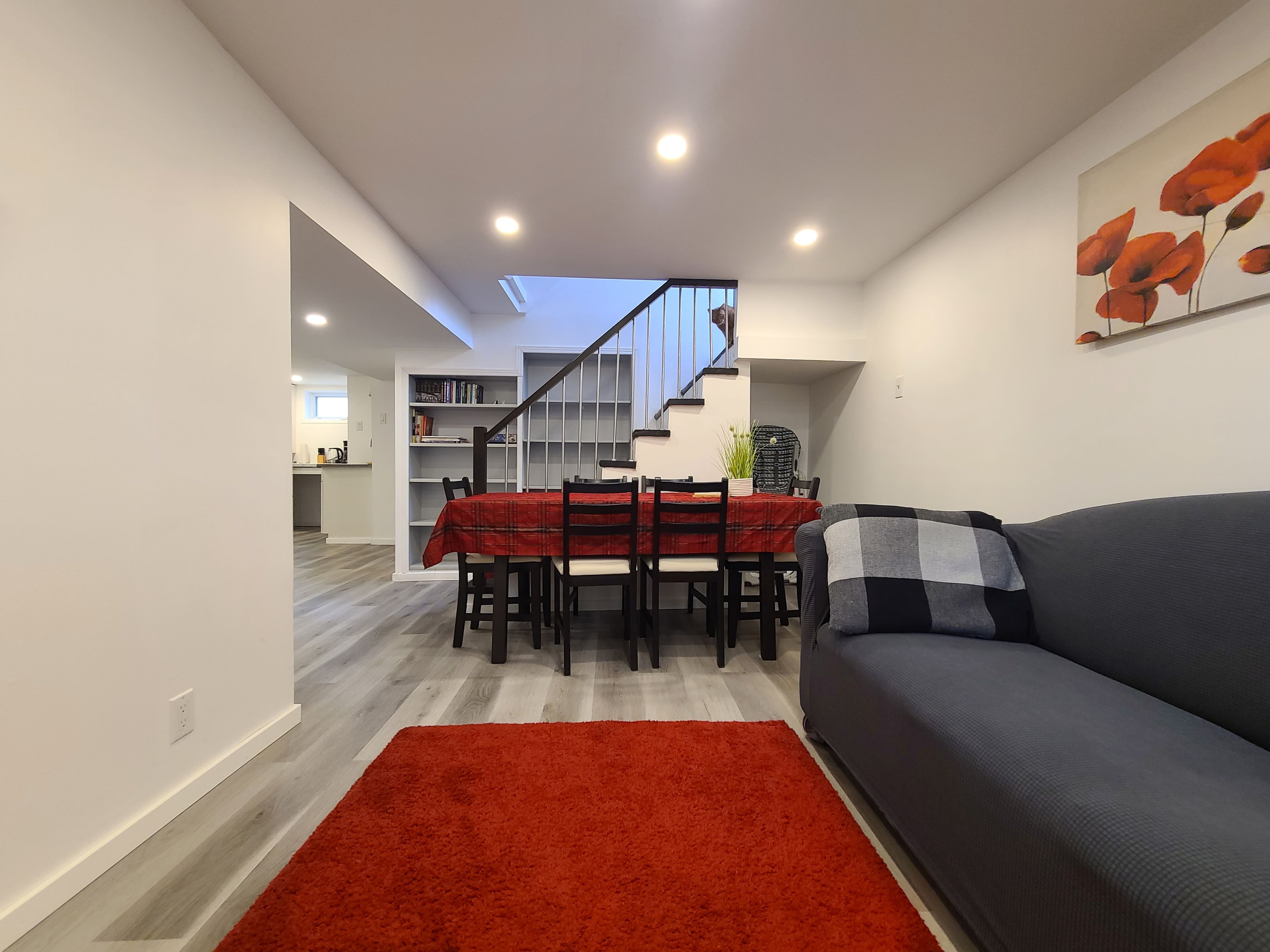 Ein modernes Wohnzimmer mit einer Couch, einem Esstisch mit Stühlen, einem roten Teppich und einer Treppe, die in eine obere Etage führt.