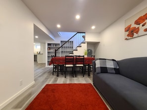 Una sala de estar moderna con un sofá, una mesa de comedor con sillas, una alfombra roja y una escalera que lleva a un nivel superior.