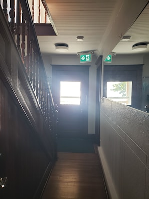 Un couloir avec un escalier en bois, une porte portant un panneau indiquant la sortie de secours et une fenêtre laissant entrer la lumière naturelle.