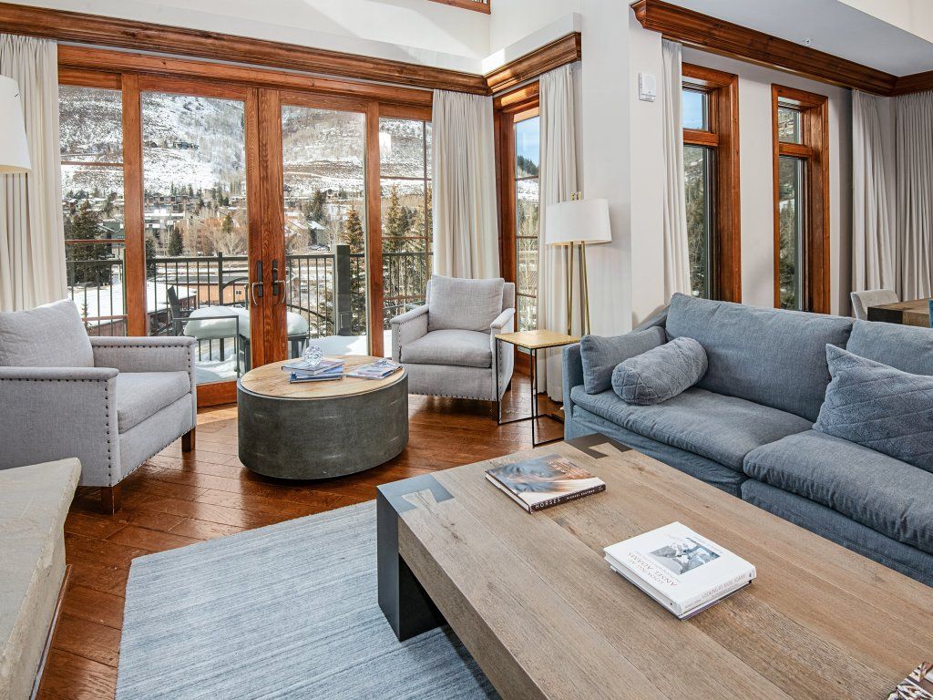 Un salon moderne avec un canapé gris, des fauteuils, une table basse et une vue sur un paysage montagneux enneigé, visible à travers de grandes fenêtres.