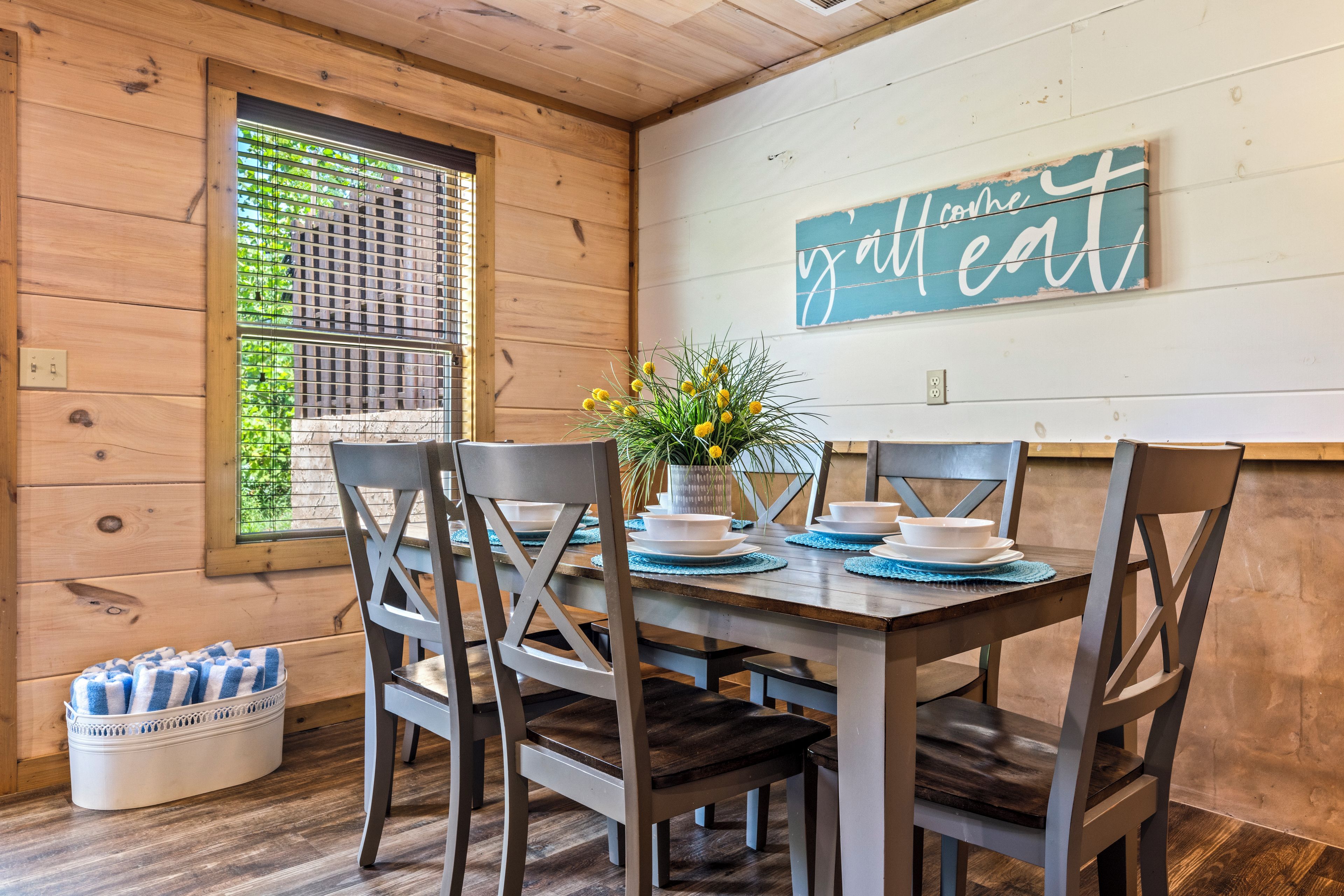 A wooden interior with a dining table set for four, surrounded by chairs, and a window with blinds.