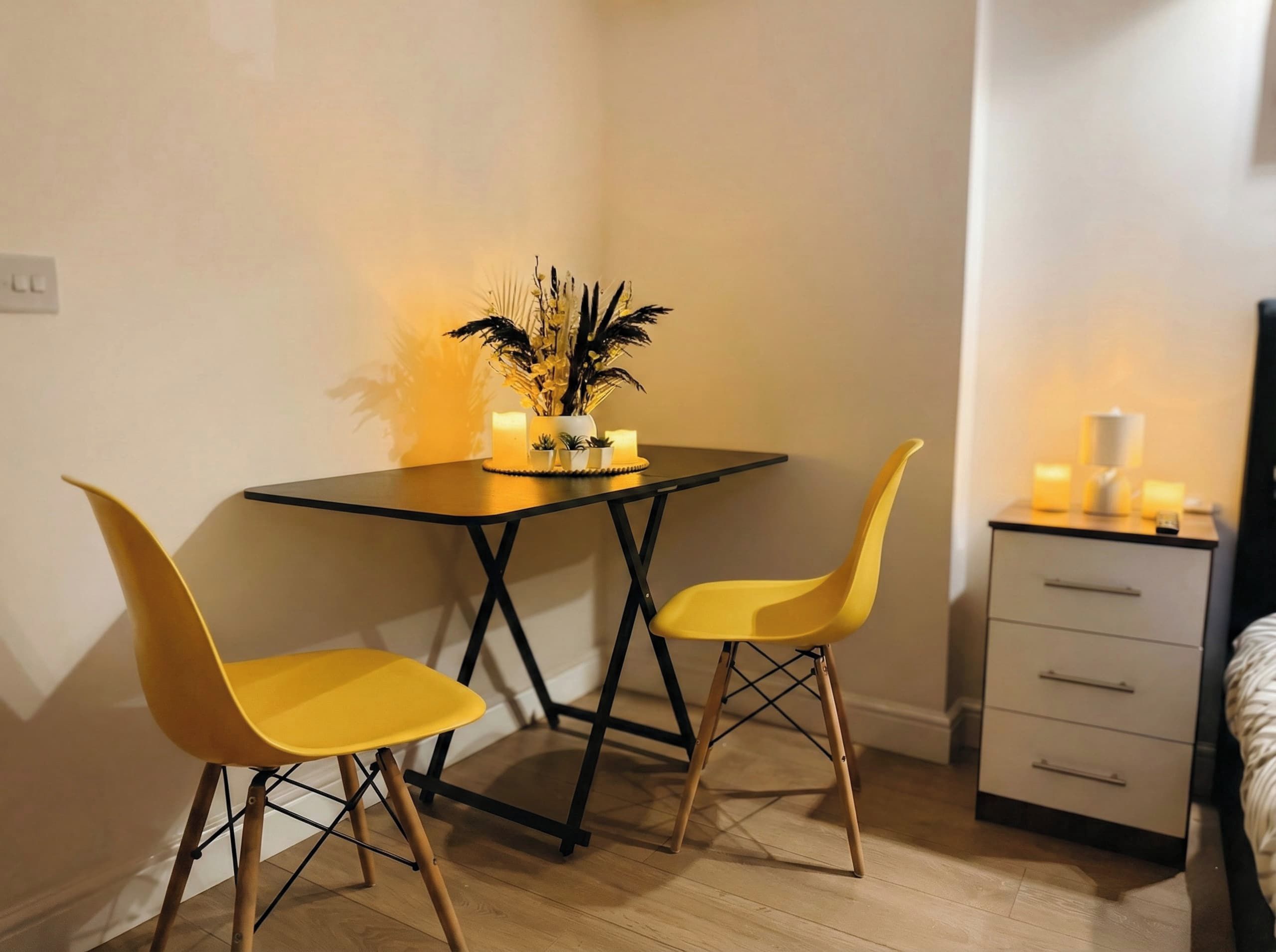 Uma pequena área de refeições com uma mesa e duas cadeiras, uma mesa de cabeceira com velas, e uma cama ao fundo.