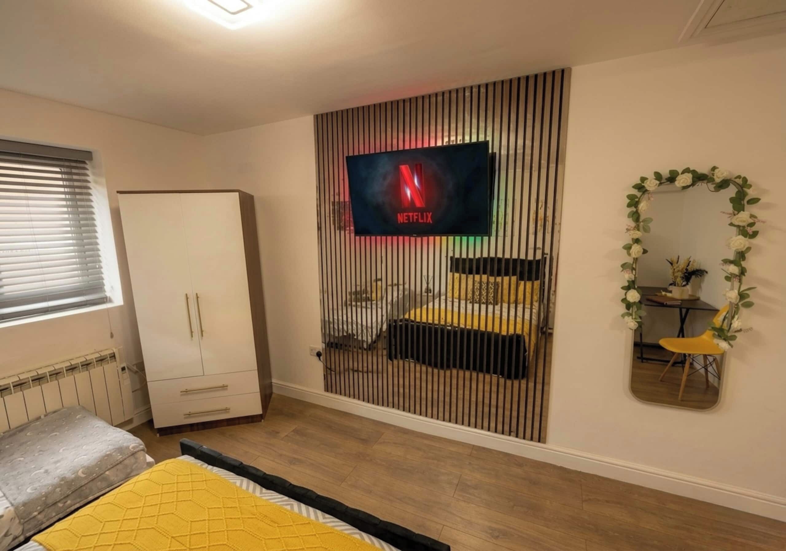 Un dormitorio con cama, armario, televisor montado en la pared y espejo con marco floral.