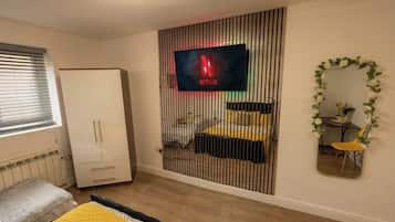 Un dormitorio con cama, armario, televisor montado en la pared y espejo con marco floral.