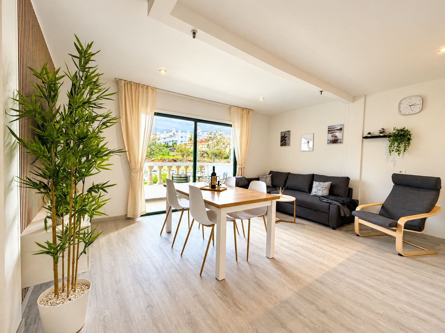 Een moderne woonkamer met een eethoek, een bank, twee fauteuils, een groot raam met uitzicht, en een klok aan de muur.