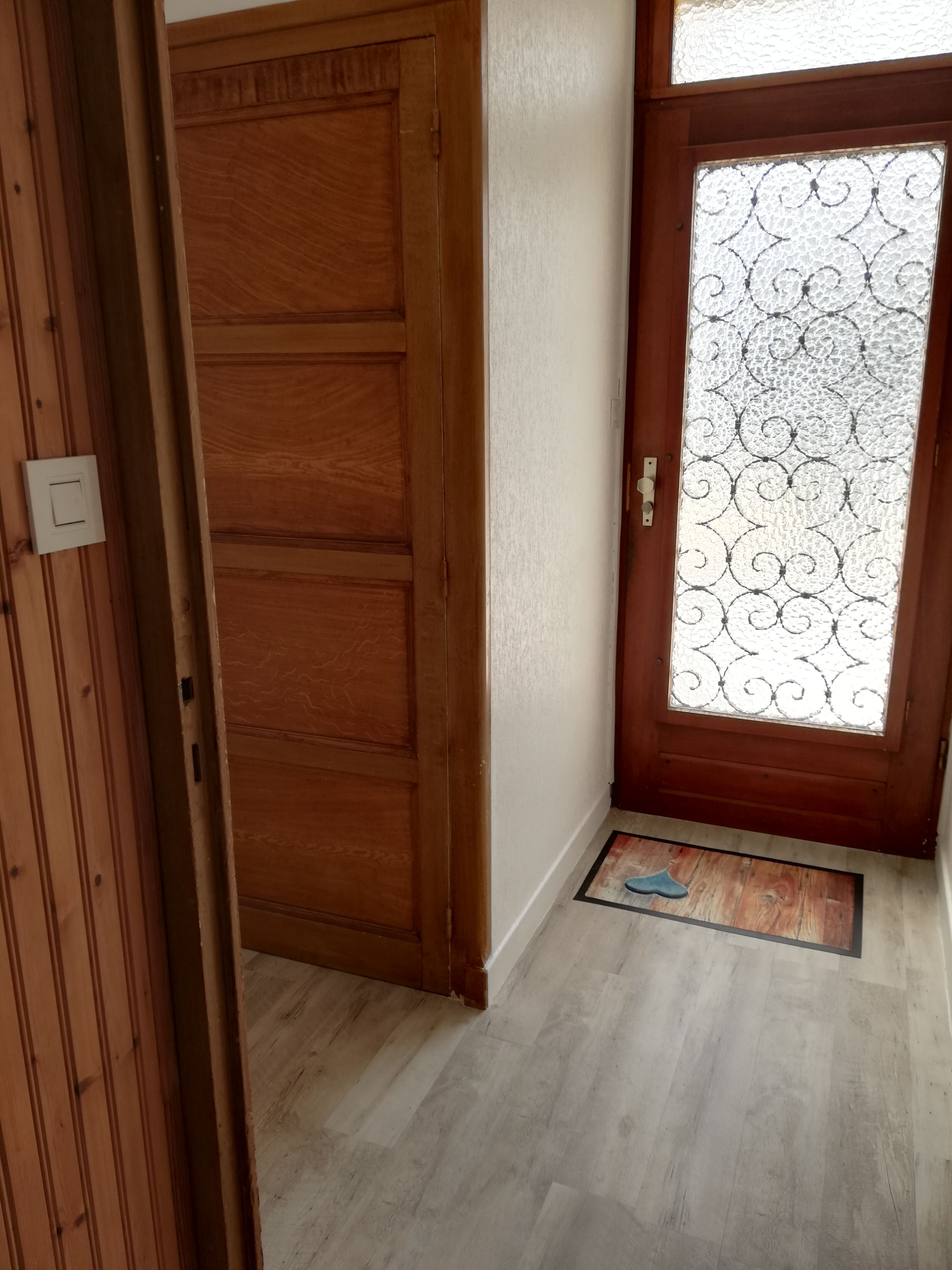 Eine Holztür mit Fensterglas, eine Fußmatte und ein Lichtschalter an der Wand.