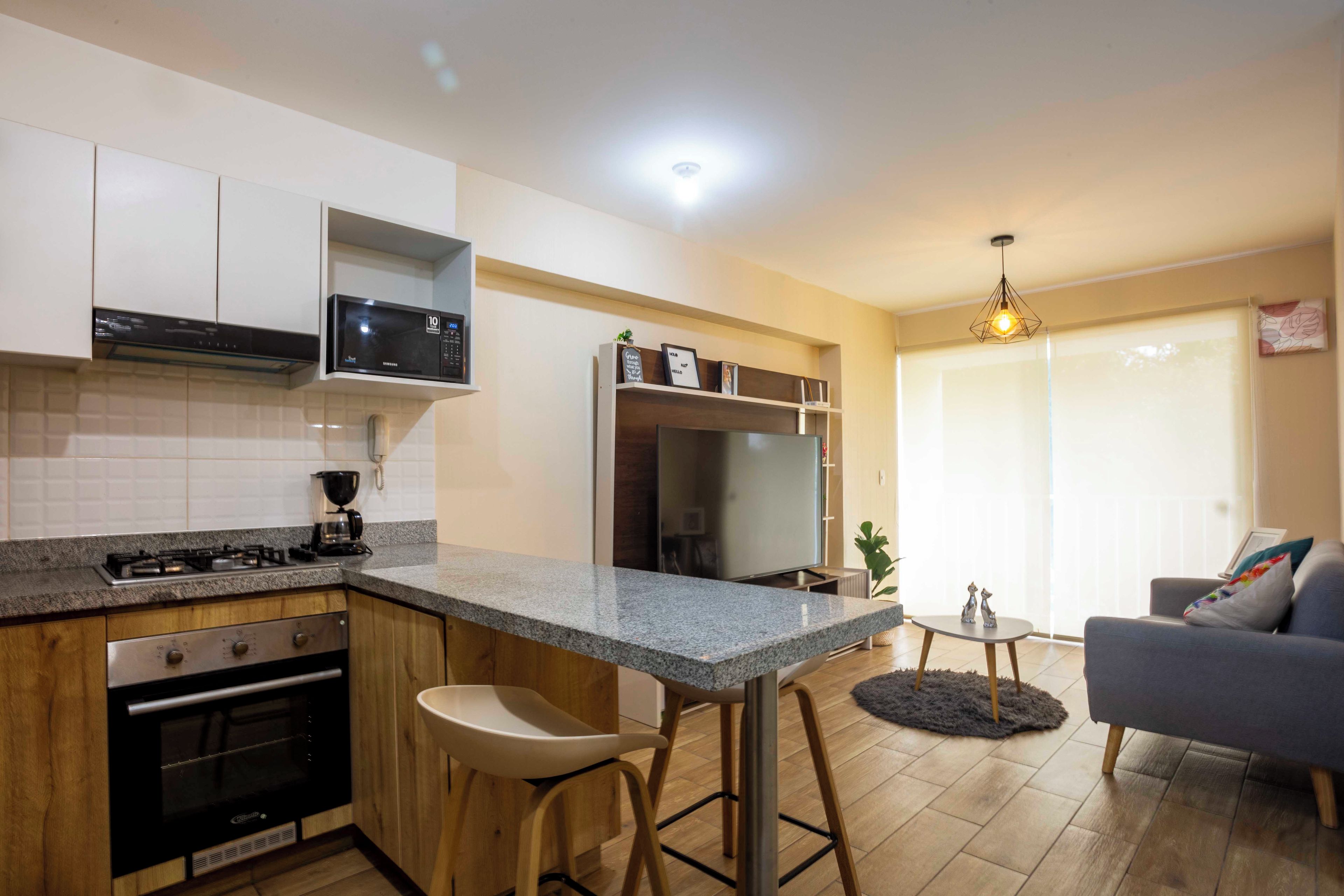 A compact kitchen with a microwave, stove, and oven. There is a bar stool and a dining area with a table and chairs.