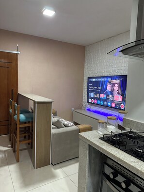 A modern kitchen with a built-in TV displaying a program, a dining area with a chair, and a small sofa. - New Listing (Angra dos Reis)