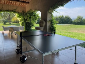 Una mesa de ping pong con red, bajo un área techada con vista a un campo de césped.