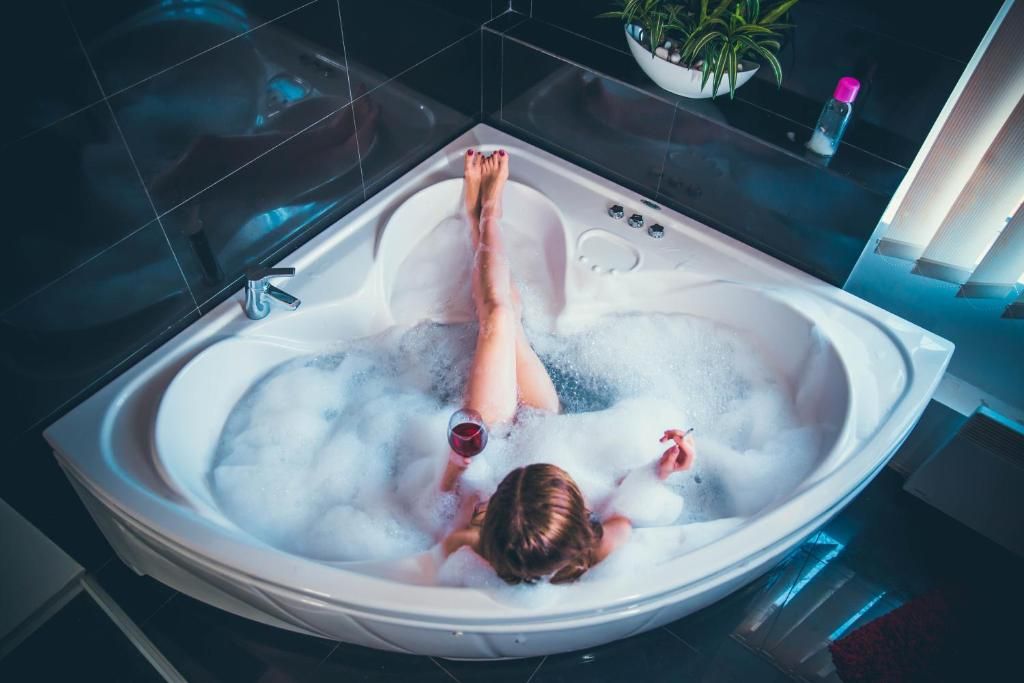 Una persona relajándose en una bañera moderna y espaciosa llena de burbujas, con una planta y una botella a un lado.