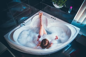 Une personne se détend dans une grande baignoire moderne remplie de mousse, avec une plante et une bouteille à proximité.