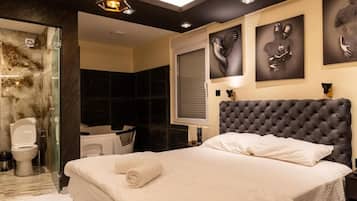 A modern hotel room with a large bed, a glass shower enclosure, a dark-toned headboard, and abstract wall art.
