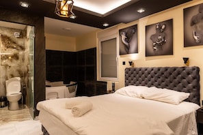 A modern hotel room with a large bed, a glass shower enclosure, a dark-toned headboard, and abstract wall art. - Vila Salute  (Niš)