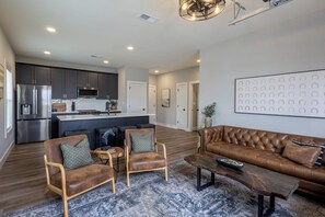 A spacious living area with a brown leather sofa, two armchairs, and a coffee table. A kitchen with dark cabinets and stainless steel appliances is visible in the background. - Bluegrass BnB - New, Downtown BG 1 Bedroom Apartment (Bowling Green)