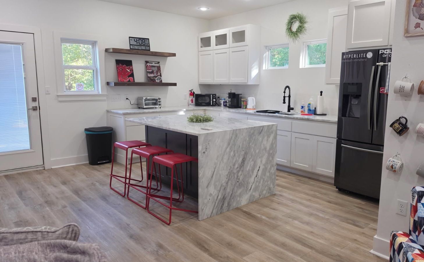Una cocina moderna con una isla de mármol, taburetes rojos, armarios blancos y un refrigerador negro.
