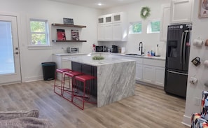 Una cocina moderna con una isla de mármol, taburetes rojos, armarios blancos y un refrigerador negro.