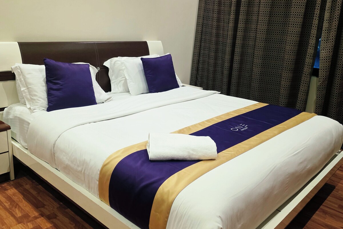 A neatly made bed with white bedding, a purple and gold accent, and a white towel on the side.