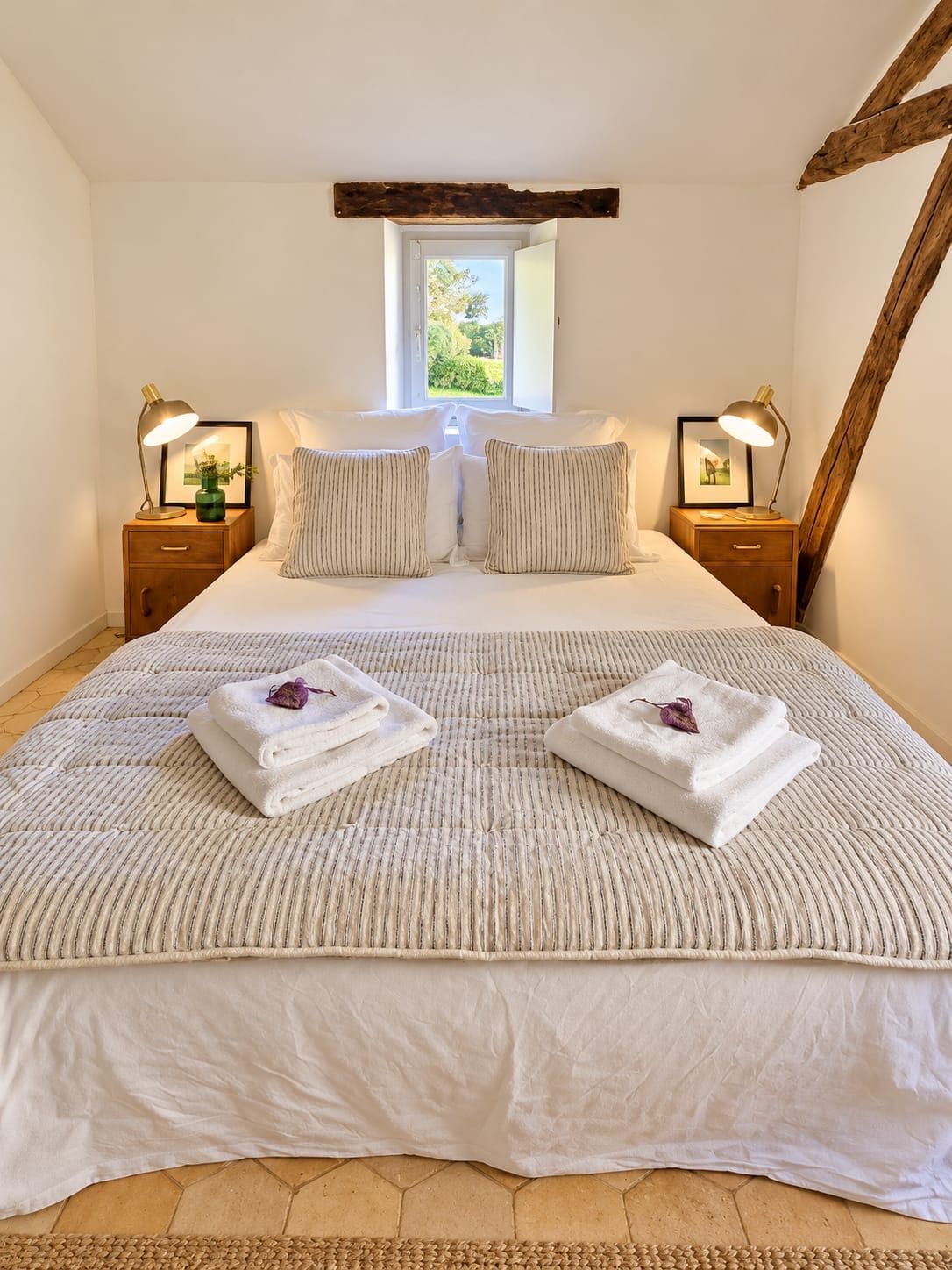 A bedroom with a large bed, two bedside tables with lamps, a window, and a wooden beam ceiling.