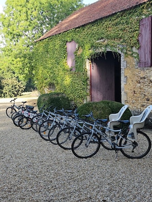 Une rangée de vélos est garée devant un bâtiment en pierre, surmonté d’un toit recouvert de tuiles rouges, et l’ensemble est entouré de verdure.