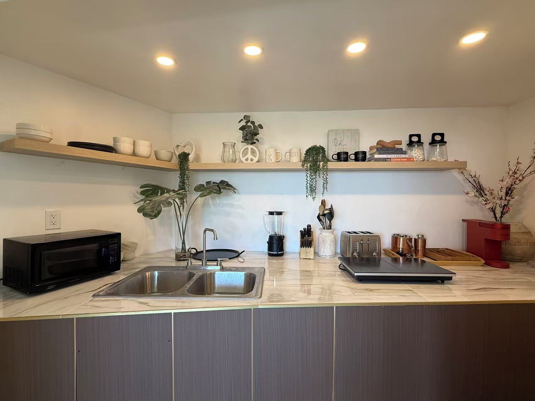 A modern kitchen with a double sink, a microwave, and a toaster. There are shelves above the counter holding various kitchen items.