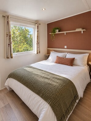 Un dormitorio ordenado con una cama, almohadas, una mesita de noche, una ventana con cortinas y un estante en la pared con una planta.