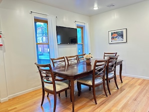 A dining area with a wooden table and chairs, a flat-screen TV mounted above the table, and a framed picture on the wall.