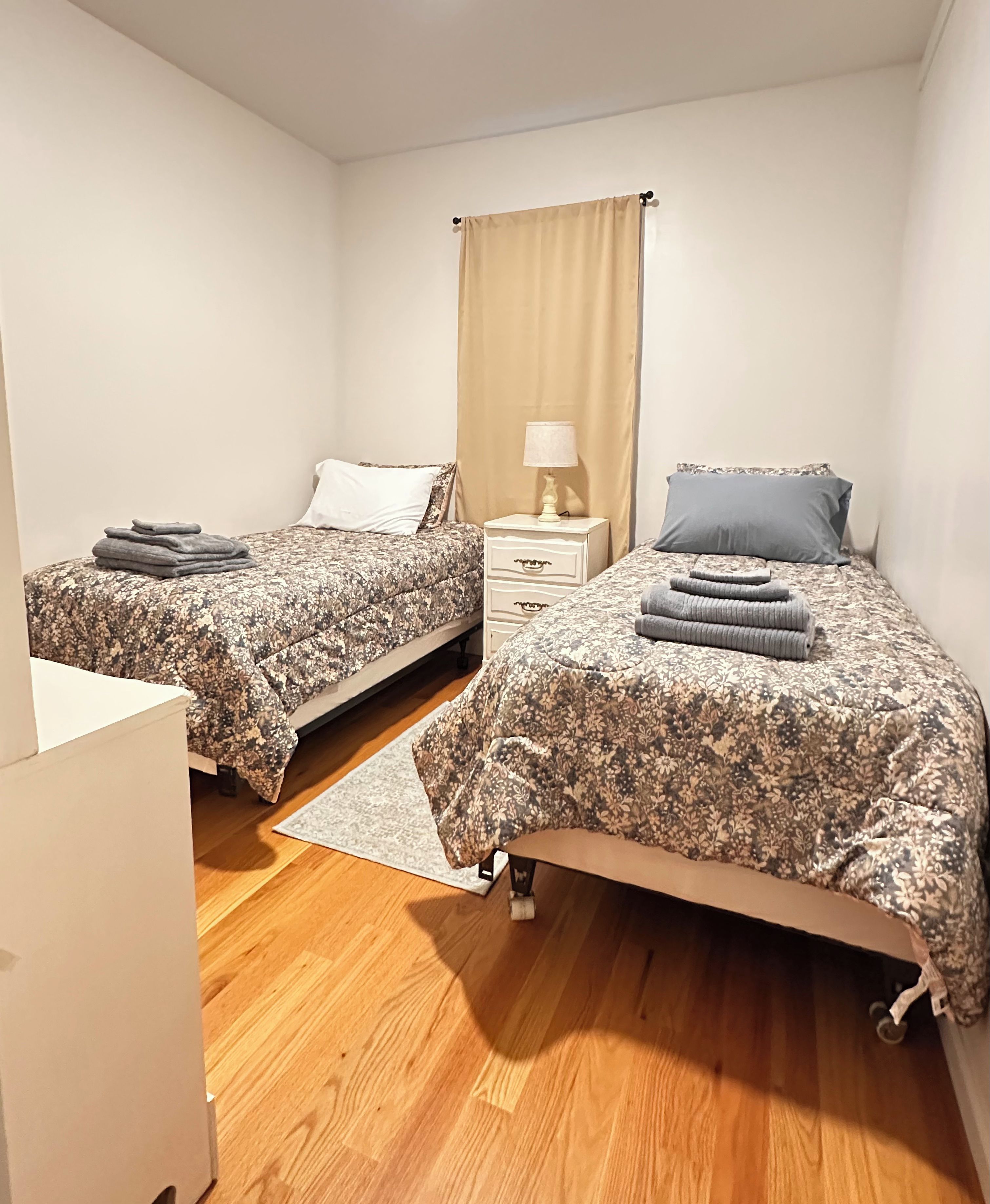 A bedroom with two beds, a nightstand, a lamp, and a window with curtains.