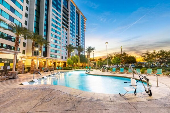Área de piscina de hotel, com uma piscina grande, espreguiçadeiras e um edifício com vários andares.