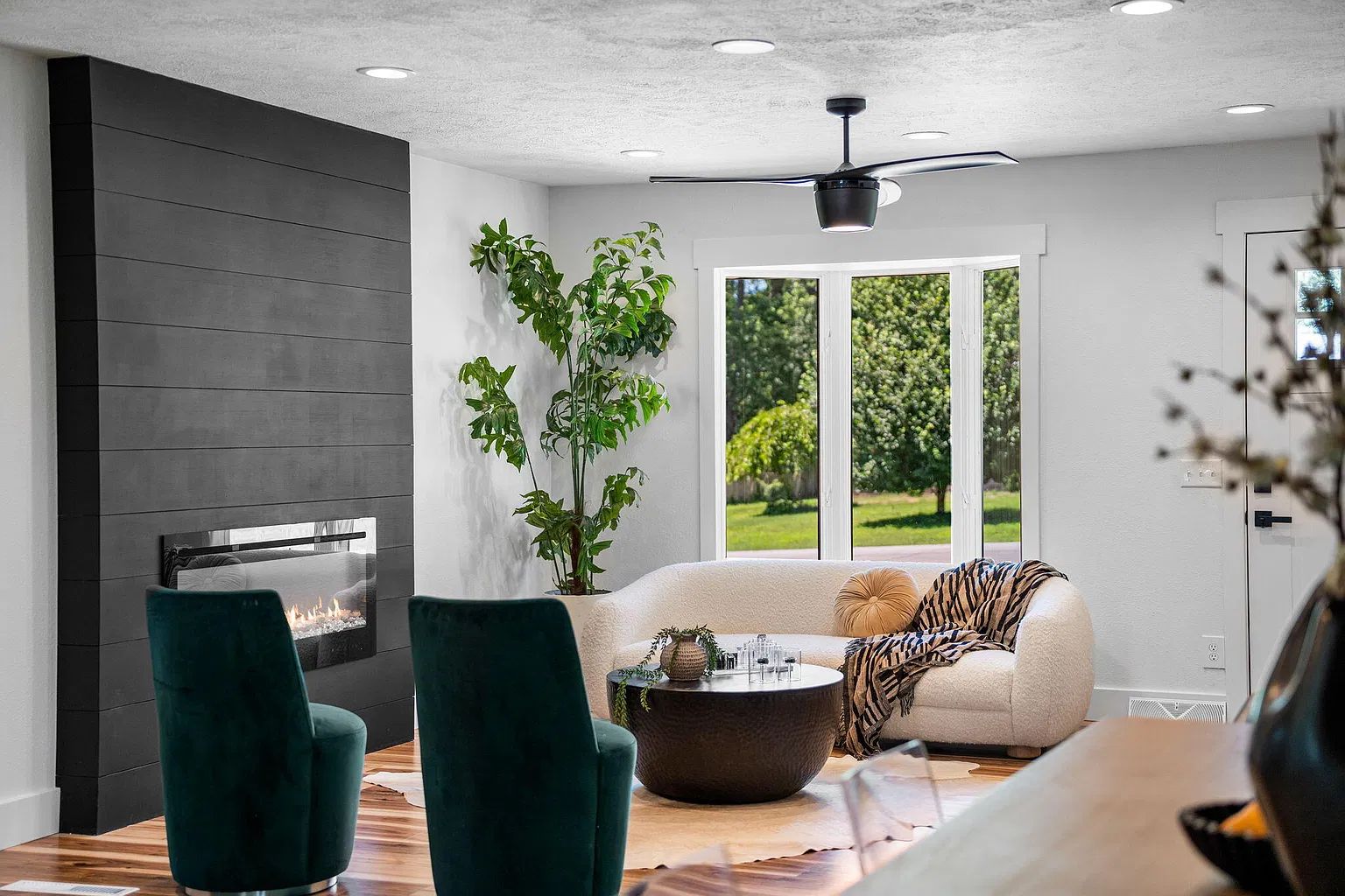 Un salon moderne avec une cheminée, des fauteuils verts, un canapé agrémenté d’un plaid à motifs zébrés et une grande fenêtre donnant sur un paysage verdoyant.