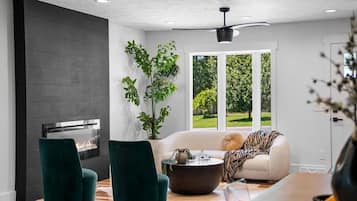 Una sala de estar moderna con chimenea, sillones verdes, un sofá con una manta de estampado de cebra y un ventanal con vistas a zonas verdes.