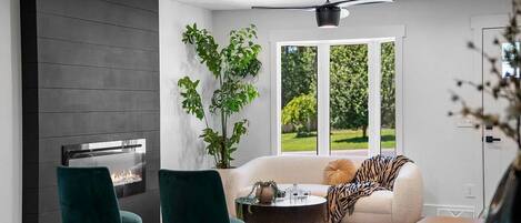 Un salon moderne avec une cheminée, des fauteuils verts, un canapé agrémenté d’un plaid à motifs zébrés et une grande fenêtre donnant sur un paysage verdoyant.