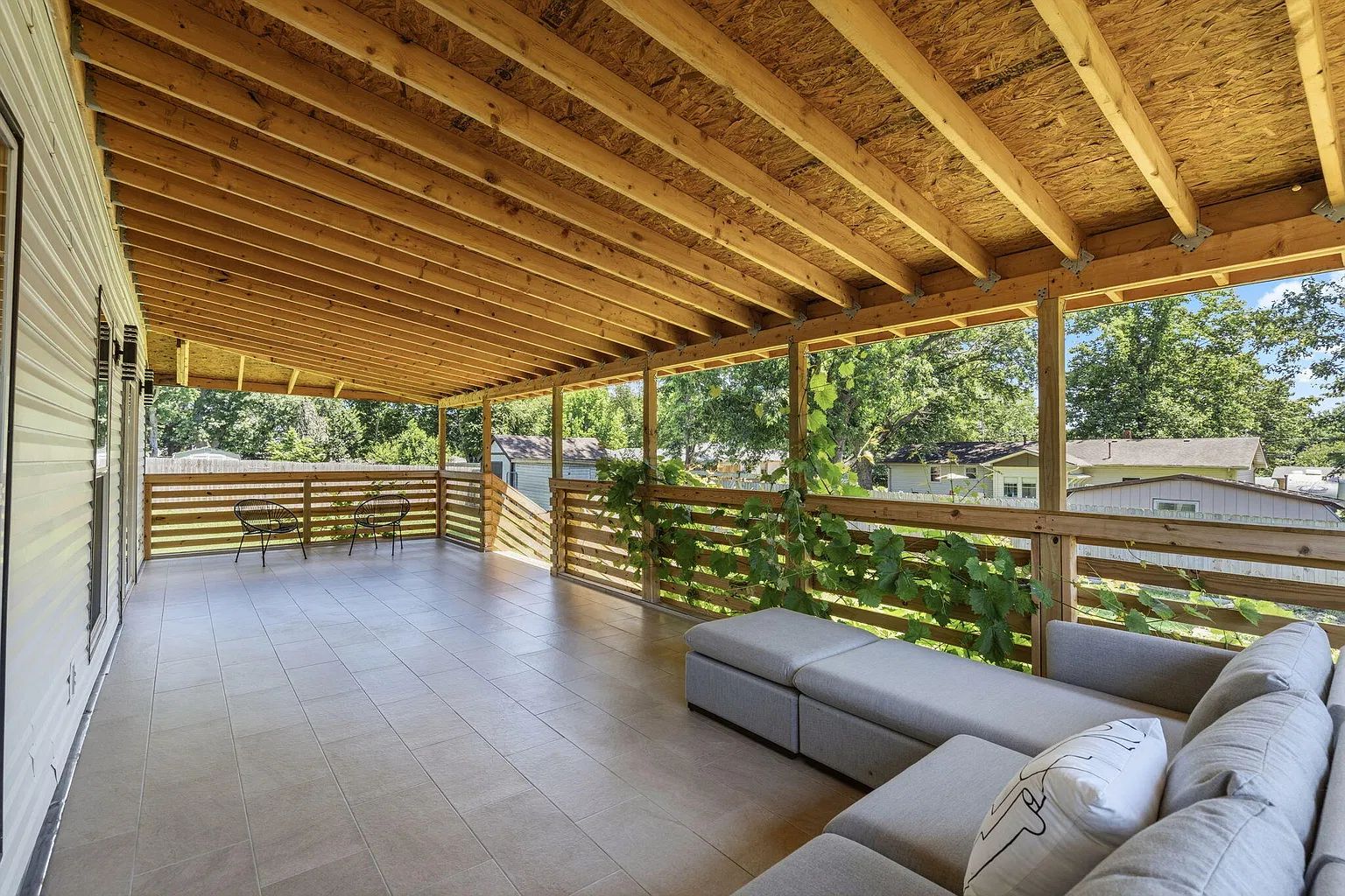 A covered outdoor patio with a wooden ceiling, a seating area with cushions, and a view of the surrounding neighborhood.