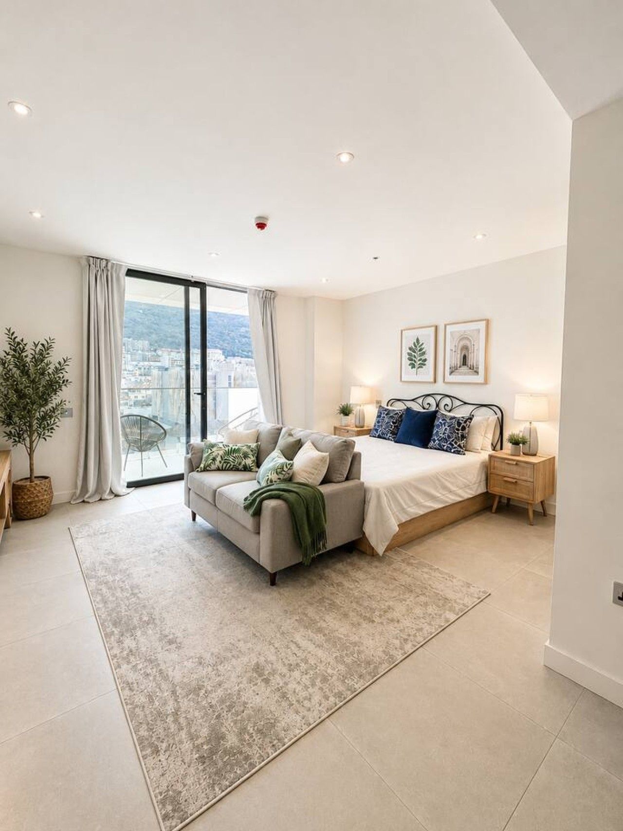 A modern bedroom with a large bed, a sofa, a bedside table, a lamp, and a view of the city through a large window.