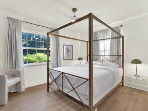 Una cama con dosel de madera, ropa de cama blanca y vistas a la vegetación a través de una ventana.