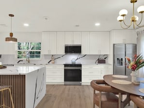 A modern kitchen with white cabinetry, a central island, stainless steel appliances, and a dining area with a wooden table and chairs. - Casa Selva (Orlando)
