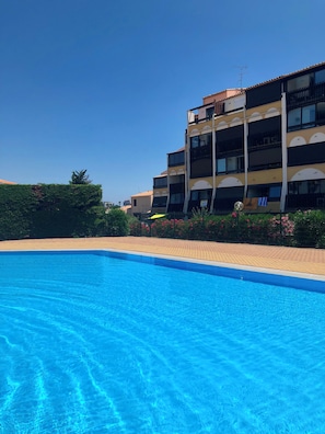Una piscina con superficie de agua azul cristalina, rodeada por un camino pavimentado y un edificio de varios pisos con balcones.