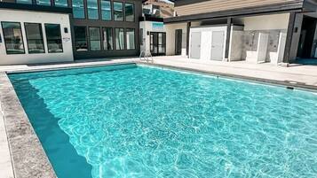 Ein Schwimmbecken mit klarem Wasser, neben einem Gebäude mit großen Fenstern und einem überdachten Bereich mit Schließfächern.