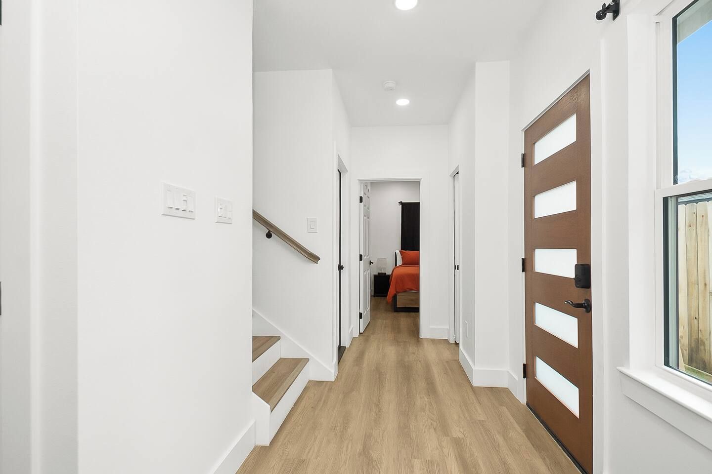 A hallway with a wooden staircase, white walls, and a door with a window.