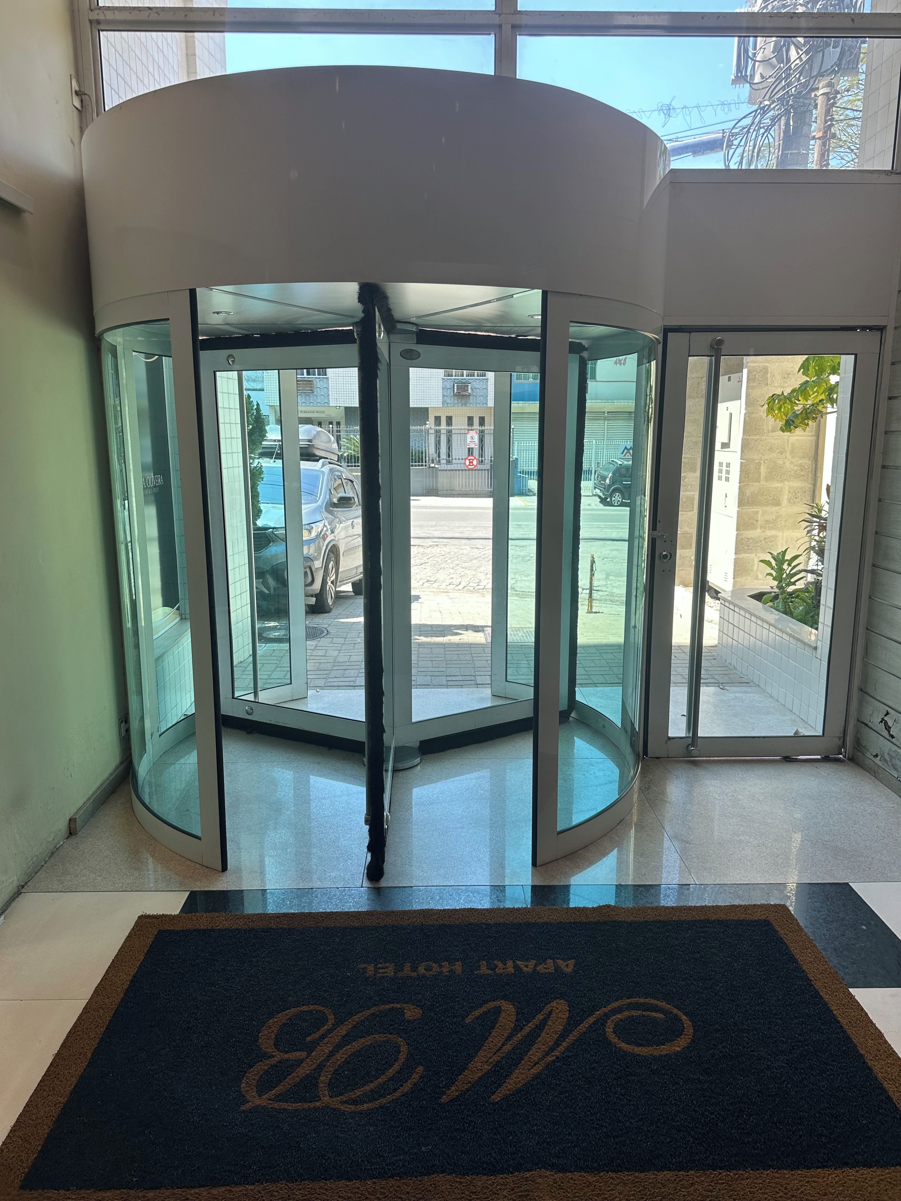 A revolving door with a glass panel, a doormat with a monogram, and a view of a parking area with a building in the background.